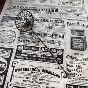 May include: A vintage-style metal skimmer with a circular wire mesh head and a long, twisted wire handle. The tool features a small loop at the end of the handle for hanging. The mesh is constructed from thin, dark-toned metal wire.