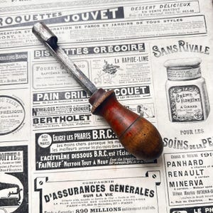 Peut inclure: Un outil en métal vintage avec une tige en acier cylindrique et patinée, dotée d'une extrémité crantée et d'un manche en bois brun foncé arrondi avec un grain visible. L'outil présente une conception robuste et utilitaire avec une poignée en bois lisse.
