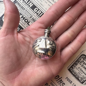 Könnte beinhalten: Eine kleine, silberfarbene Parfümflasche mit einem katzenartigen Gesichtsdesign. Die Flasche hat eine runde Form und einen kleinen, grünen Verschluss. Die Flasche wird in einer Hand gehalten, mit einer Vintage-Zeitung im Hintergrund.