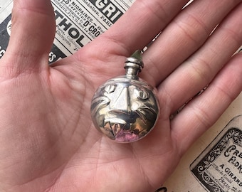 Puede incluir: Un pequeño frasco de perfume plateado con un diseño de cara de gato. El frasco tiene forma redonda y un pequeño tapón verde. El frasco se sostiene en una mano, con un periódico antiguo en el fondo.