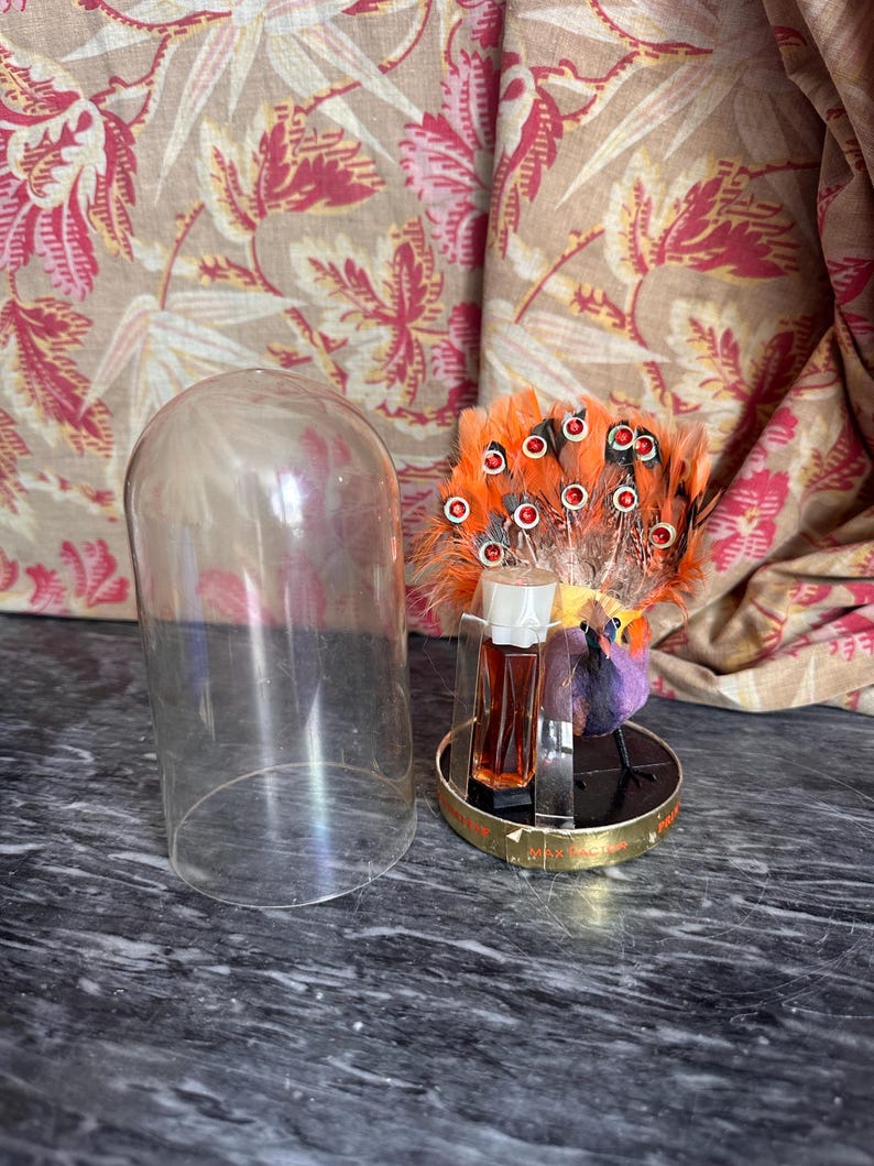 Puede incluir: Un frasco de perfume vintage Max Factor con una figura decorativa de p&aacute;jaro con plumas de color naranja brillante y detalles de pedrer&iacute;a roja. El p&aacute;jaro y el peque&ntilde;o frasco de vidrio rectangular con tapa blanca est&aacute;n montados sobre una base dorada circular, acompa&ntilde;ados por una c&uacute;pula de vidrio transparente.
