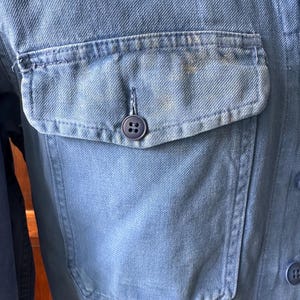 May include: A light blue denim shirt featuring a single chest pocket with a flap and a dark blue button. The fabric displays a visible twill weave texture and subtle fading throughout the material. Additional dark blue buttons are visible.