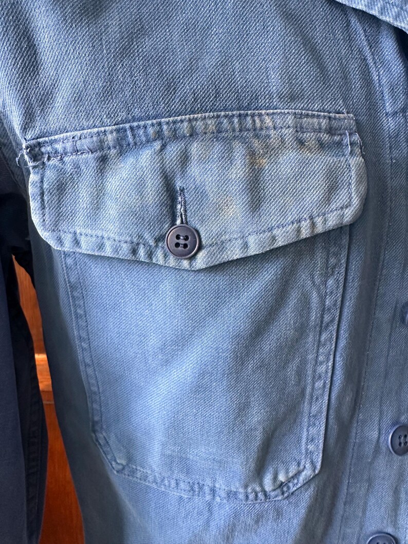May include: A light blue denim shirt featuring a single chest pocket with a flap and a dark blue button. The fabric displays a visible twill weave texture and subtle fading throughout the material. Additional dark blue buttons are visible.