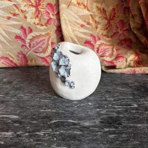 Op de afbeelding: Een kleine, gebroken witte, ronde keramische vaas met een gladde, matte afwerking. Een cluster van kleine, metalen, zilverkleurige bloemvormige versieringen is bevestigd aan de linkerbovenkant van de vaas, met gedetailleerde bloemblaadjes.