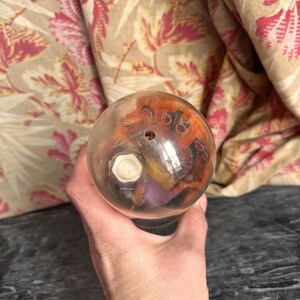 Puede incluir: Un pisapapeles esf&eacute;rico de cristal transparente que contiene una peque&ntilde;a criatura naranja y negra con ojos visibles y un componente blanco en forma de perno hexagonal incrustado en el centro de la estructura del orbe.