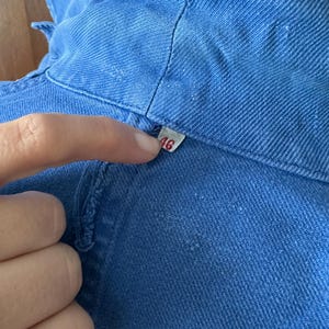 May include: A blue cotton-twill work jacket featuring a visible collar and a small white interior tag printed with the number 46. The garment displays a textured weave pattern and a structured neckline with visible stitching along the seams.