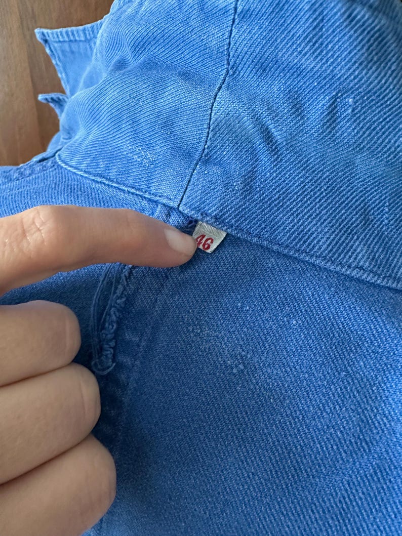 May include: A blue cotton-twill work jacket featuring a visible collar and a small white interior tag printed with the number 46. The garment displays a textured weave pattern and a structured neckline with visible stitching along the seams.