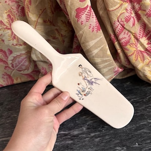 Op de afbeelding: Off-white keramische taartschep met een handvat en een breed, plat blad. Het blad heeft een vintage illustratie van mensen in formele kleding. De schep wordt door een hand vastgehouden.