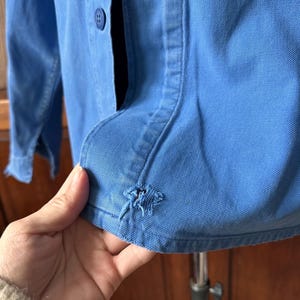 May include: A bright blue long-sleeved button-down shirt featuring a visible frayed hole near the bottom hem. The garment is constructed from a sturdy twill fabric and includes a matching blue plastic button on the front placket.
