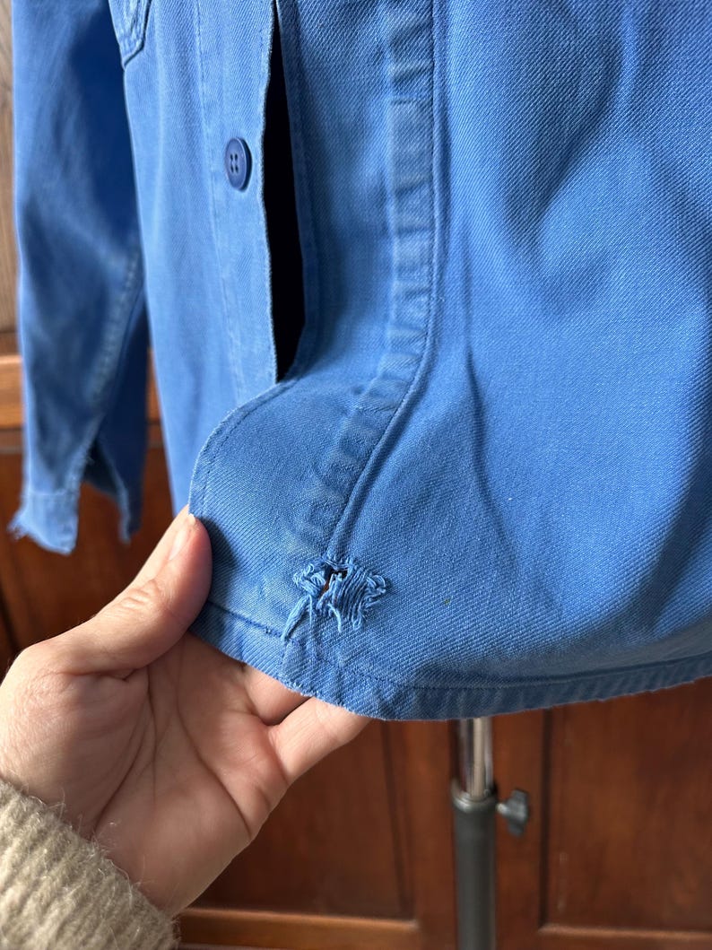 May include: A bright blue long-sleeved button-down shirt featuring a visible frayed hole near the bottom hem. The garment is constructed from a sturdy twill fabric and includes a matching blue plastic button on the front placket.
