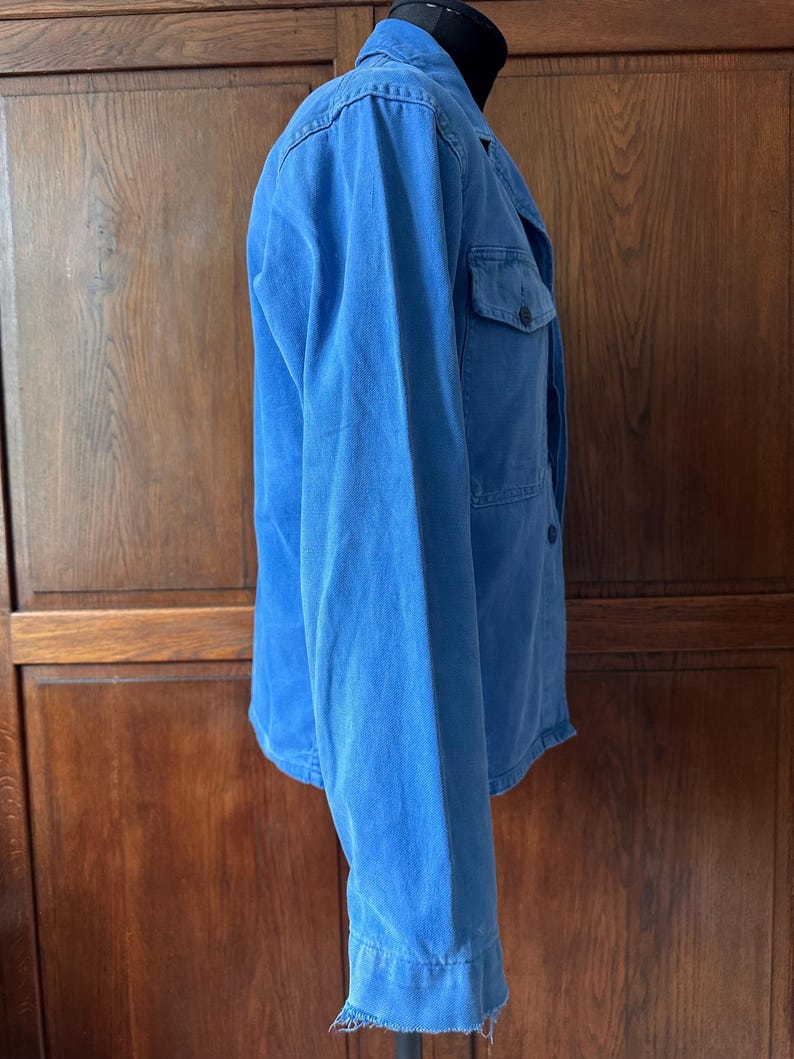 May include: A bright blue long-sleeved jacket featuring a collared neckline, a button-down front, and a single chest pocket. The garment is displayed on a mannequin and has a raw, frayed hemline at the bottom of the sleeves and the torso.