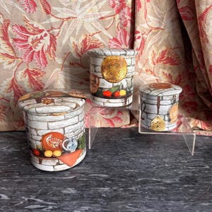 May include: Three cylindrical metal canisters with lids, each featuring a printed design of a white brick wall, kitchen utensils, and various food items like tomatoes and lemons. The canisters vary in size and are arranged on a clear display stand.