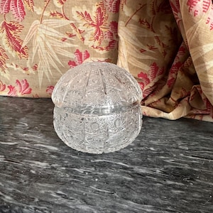 Könnte beinhalten: Ein klarer, geschliffener Glasbehälter mit gewölbtem Deckel. Der Behälter ist rund mit einem detaillierten, geometrischen Muster. Der Hintergrund zeigt gemusterte Stoffe in Rot-, Beige- und Brauntönen und eine dunkelgraue Marmoroberfläche.