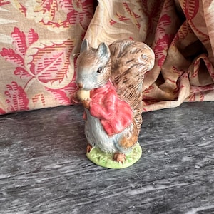 Op de afbeelding: Een keramieken eekhoornbeeldje, gekleed in een rode jas en met een noot. De eekhoorn heeft een bruin en wit lichaam, een volle staart en staat op een groene basis. De achtergrond heeft een bloemmotief.
