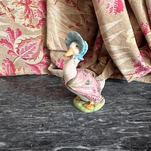 Op de afbeelding: Een keramieken eendenbeeldje met een blauwe hoed en een roze jurk met groene bloemaccenten. De eend heeft een gele snavel en staat op een groene basis. De achtergrond bevat een bloemmotiefstof.
