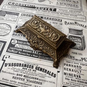 May include: A rectangular brass inkwell featuring an ornate, raised relief design of scrolling acanthus leaves and floral motifs. The object has a hinged lid with a central decorative panel and stands on four small, curved feet with intricate detailing.