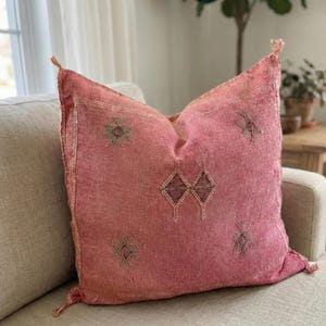 May include: A square, pink decorative pillow with embroidered geometric patterns. The pillow has a textured appearance and is adorned with tassels at each corner. The pillow is resting on a beige sofa.