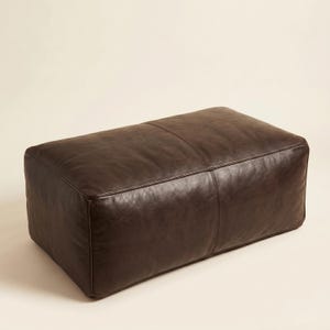 May include: A rectangular, dark brown leather ottoman. The ottoman has a stitched seam detail running down the center and along the edges. The leather has a slightly textured appearance.