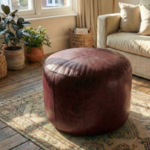 May include: A round, burgundy leather ottoman sits on a patterned rug in a living room setting. The ottoman is the focal point, with visible stitching and a slightly distressed appearance. Plants and a sofa are in the background.