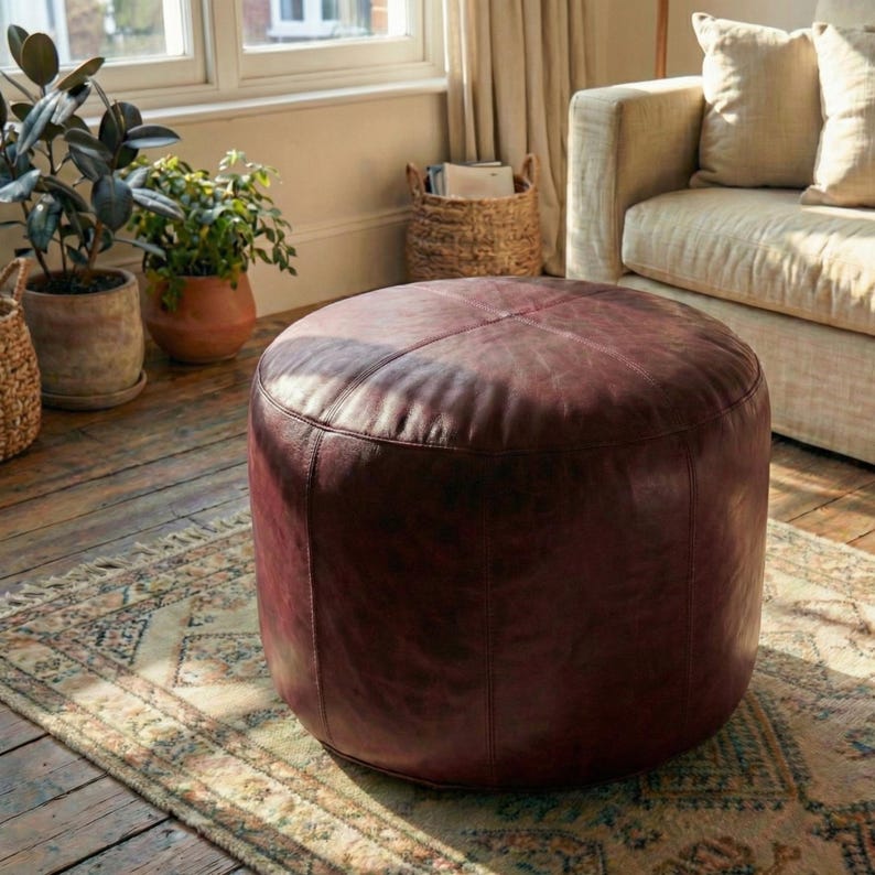 May include: A round, burgundy leather ottoman sits on a patterned rug in a living room setting. The ottoman is the focal point, with visible stitching and a slightly distressed appearance. Plants and a sofa are in the background.