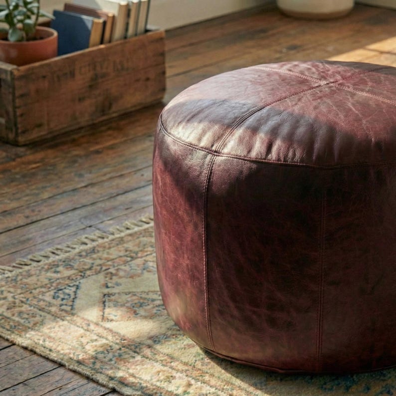 May include: A round, burgundy leather ottoman with stitched seams. The ottoman is placed on a wooden floor next to a patterned rug. The leather has a distressed appearance.