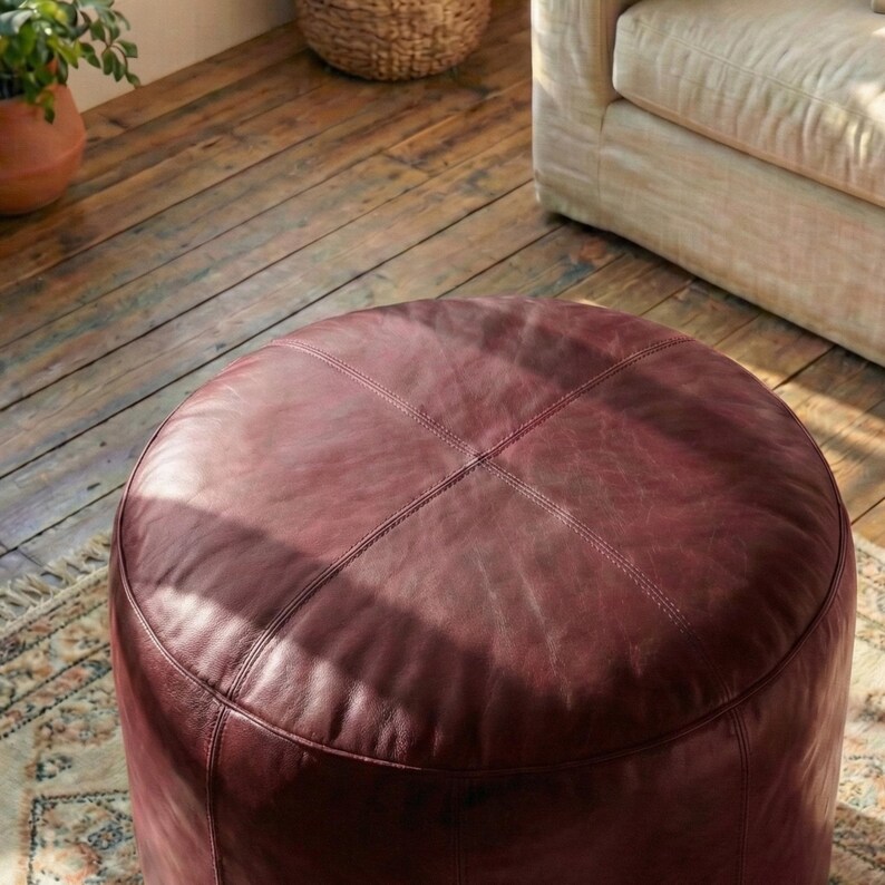 May include: A round, burgundy leather ottoman with stitched detailing. The ottoman is a deep reddish-brown color and has a smooth, slightly textured surface. It is a piece of furniture, likely for a living room or similar space.
