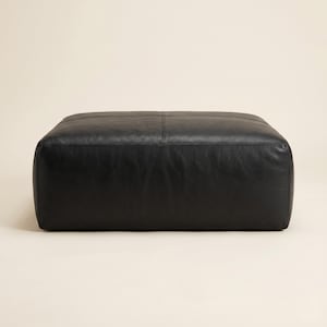 May include: A large, square black leather ottoman. The ottoman has a stitched seam detail across the top and is made of a smooth, solid black leather. The design is simple and modern, suitable for a living room or lounge area.