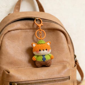 Pode incluir: Um chaveiro de raposa de crochê, laranja com detalhes brancos, um chapéu e camisa verdes, uma gravata azul e calças marrons. Ele está pendurado em um anel e fecho de metal laranja. O fundo é uma mochila de couro marrom.