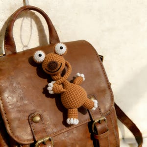 May include: A brown crocheted creature with large white-rimmed eyes and white paws is attached to a brown leather bag. The handmade toy has a smiling face and is made of a soft, textured yarn. The bag has a buckle and a handle.