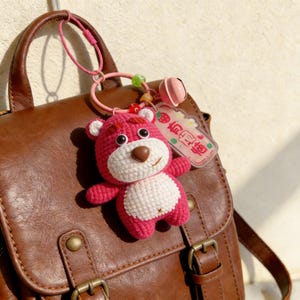 May include: A pink crocheted bear keychain with a white belly and a brown nose. The bear has black eyes and white accents on its ears. It hangs from a pink ring attached to a brown leather backpack. A small pink bell and a tag with Japanese text are also attached.