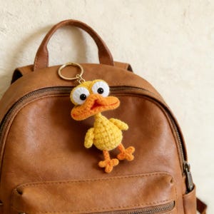 Puede incluir: Un llavero de pato de ganchillo amarillo y naranja hecho a mano con grandes ojos saltones. El pato tiene pico, patas y pequeñas alas naranjas. El llavero está sujeto a un anillo dorado y cuelga de una mochila de cuero marrón.
