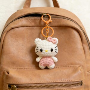 May include: A handmade crochet keychain featuring a white cat with a pink bow and overalls. The cat has black eyes, whiskers, and a yellow nose. The keychain is attached to an orange clasp and a brown backpack.