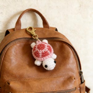 May include: A crocheted turtle keyring in white, pink, and red, attached to a gold-coloured clasp. The turtle is hanging on a brown leather backpack. The turtle's shell has a patterned design.