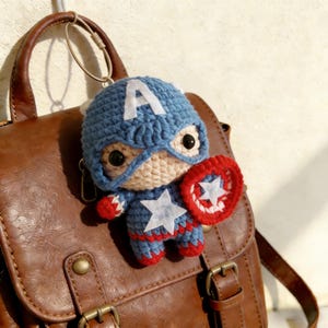 Op de afbeelding: Een handgemaakte Captain America sleutelhanger. Het gehaakte figuur heeft een blauwe helm met een witte "A", een masker en een schild. Het figuur is blauw, rood en wit en is bevestigd aan een zilveren sleutelring. De sleutelhanger hangt aan een bruine leren tas.