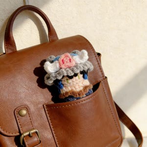 May include: A handmade crochet doll with a floral headpiece peeks out of a brown leather bag pocket. The doll features a beige face, blue hair, and a gray and white hat with a pink rose. The bag has a buckle closure.