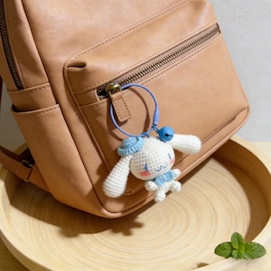 May include: A white crochet keychain in the shape of a cartoon character with long ears, a blue hat, and a blue bow tie. The keychain is attached to a tan backpack with a blue loop and a small blue bell.