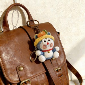 May include: A handmade crochet keychain featuring a blue and white cartoon cat wearing a yellow hat with a carrot on top. The keychain has a red collar with a small gold bell and is attached to a brown leather backpack.