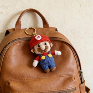 Può includere: Portachiavi di Mario all'uncinetto con cappello rosso con una "M" bianca, baffi marroni e tuta blu. Il portachiavi è attaccato a un anello color oro ed è esposto su uno zaino in pelle marrone.