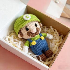 May include: A handmade crochet doll of a character wearing a green hat with a white emblem, a brown moustache, and blue dungarees with yellow buttons. The doll is in a white box with brown paper filler.