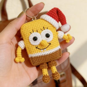May include: A handmade yellow crochet keychain featuring a cartoon character wearing a red and white Santa hat. The character has large white eyes, a smiling face, and brown shorts. The keychain is attached to a silver ring.