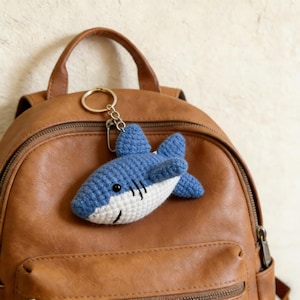 Könnte beinhalten: Ein blau-weißer gehäkelter Hai-Schlüsselanhänger mit silbernem Verschluss und Ring. Der Hai hat schwarze Details für Augen und Kiemen. Der Schlüsselanhänger ist an einem braunen Lederrucksack befestigt.