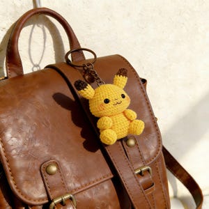 May include: A yellow crocheted Pikachu keyring with brown-tipped ears and black eyes. The plush toy is attached to a bronze-coloured keyring and is hanging on a brown leather backpack. The backpack has a buckle closure and a top handle.
