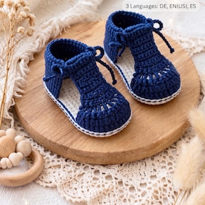 Könnte beinhalten: Handgefertigte, marineblaue Baby-Häkelsandalen mit weißen Sohlen und zarten Bindebändern. Sie liegen auf einem Holzbrett, im Hintergrund ein cremefarbenes Spitzentuch. Der Text lautet "3 Languages: DE, EN(US), ES".