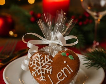 Può includere: Un biscotto di pan di zenzero a forma di cuore decorato con glassa bianca raffigurante un fiocco di neve e il nome Leni, sormontato da una piccola foglia di agrifoglio verde e una bacca rossa, confezionato in un sacchetto trasparente.