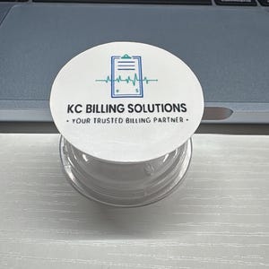 May include: A clear plastic container with a white lid featuring the text "KC BILLING SOLUTIONS - YOUR TRUSTED BILLING PARTNER." The lid has a teal and blue graphic of a clipboard and a heartbeat line. The container sits on a white surface.