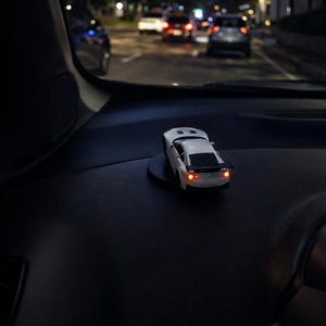 Könnte beinhalten: Ein weißes Spielzeugauto mit roten Rücklichtern steht auf einem schwarzen Armaturenbrett. Das Miniaturauto hat detaillierte Merkmale. Der Hintergrund zeigt eine nächtliche Straßenszene mit Autoscheinwerfern.