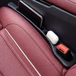 May include: A car seat organizer in black with a burgundy leather seat. A smartphone, white earbuds, and a red seatbelt buckle are visible in the organizer's compartments. The seat has a white accent stripe.