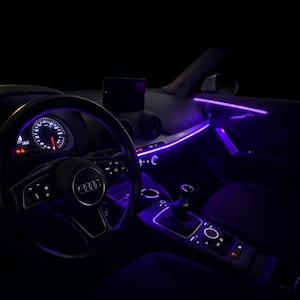 Op de afbeelding: Interieur van een auto met paarse sfeerverlichting die het dashboard en de cabine accentueert. Het Audi-logo staat op het stuurwiel en het instrumentenpaneel toont meters. De versnellingspook en middenconsole zijn ook zichtbaar.