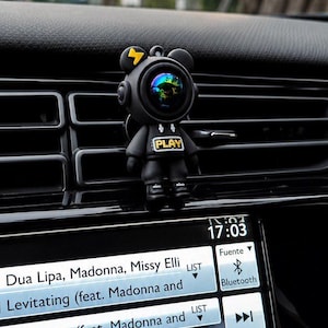 Puede incluir: Decoración negra para la ventilación del coche con forma de astronauta oso con un rayo amarillo en la cabeza. La palabra "PLAY" está en el pecho. La pantalla muestra información musical.