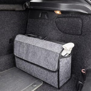 May include: A gray felt car trunk organizer with a black trim and handle, placed inside a car trunk. The organizer is rectangular with a flap closure and contains white items. A gray car mat is visible.
